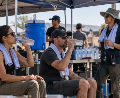 Film crew members drinking water in a shaded hydration station between outdoor takes